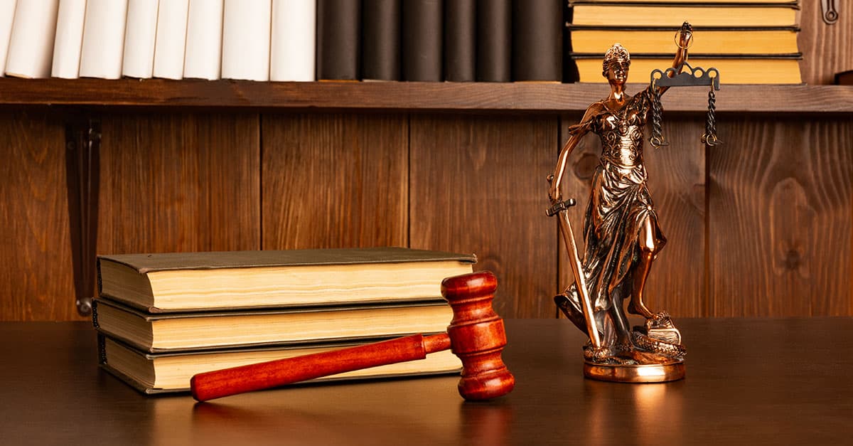 Como capa da matéria “Livros para advogados – Parte 2”, temos livros sobre uma mesa com um martelo de juiz e uma estátua da Justiça.